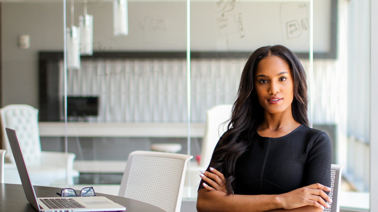 Businesswoman sitting at a desk, engaging directly with the viewer, with an open laptop on the desk in front of her, representing the professionalism and reliability of the best medical spa software of 2024